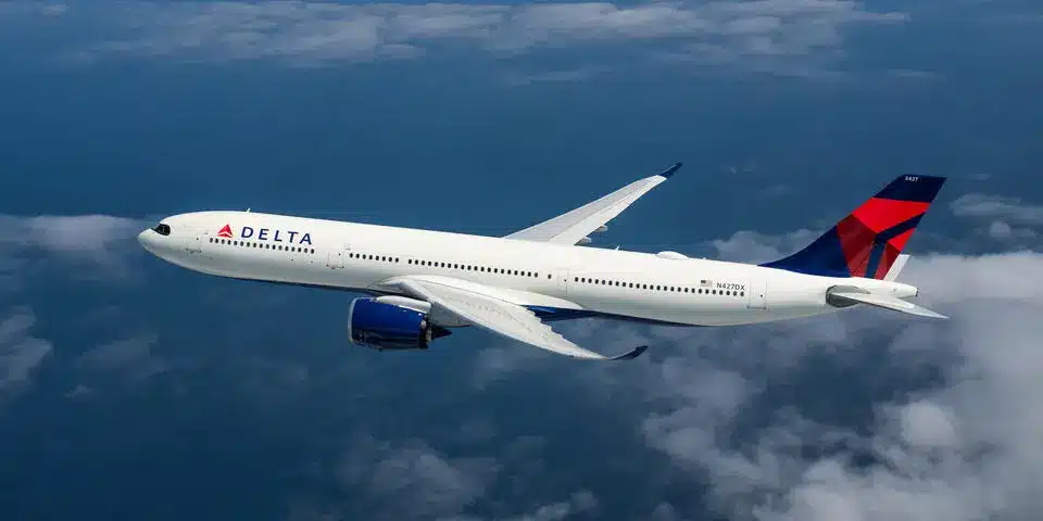 A Delta Airlines passenger jet, primarily white with blue and red tail fin colors, flies through a blue sky with scattered white clouds below and around it. The plane is seen from a slightly elevated side angle, showcasing its sleek design and two engines under its wings. The Delta logo is visible on the fuselage.